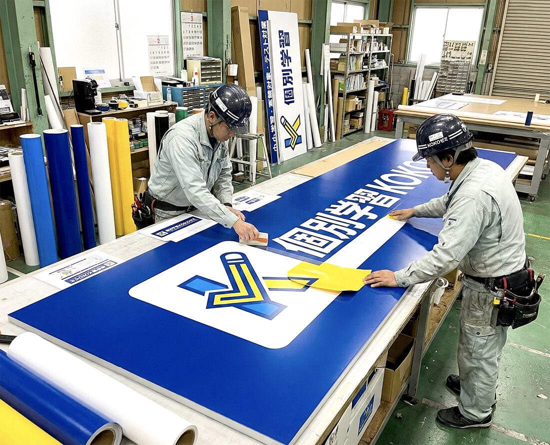 塾の看板製作中の工場内写真｜職人2名がアルミ複合パネルにシート貼り作業中