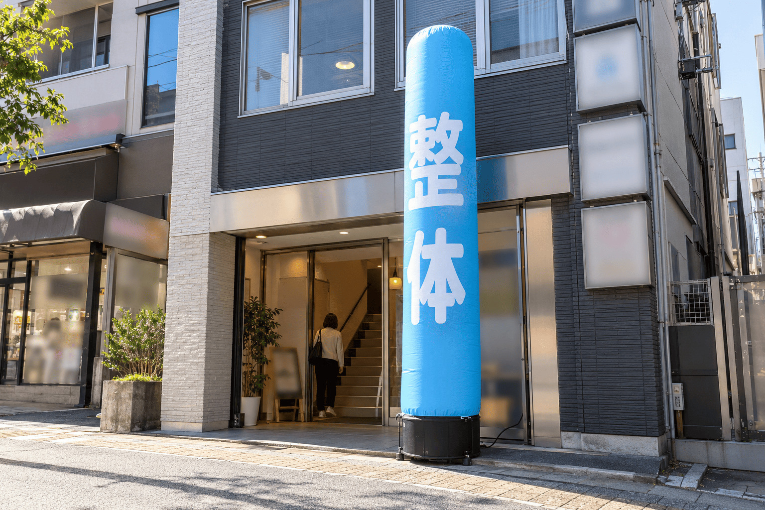 雑居ビルの1階の入口の横に、水色の地に白い文字で「整体」と書かれた全高3mのエアー看板が置かれている様子
