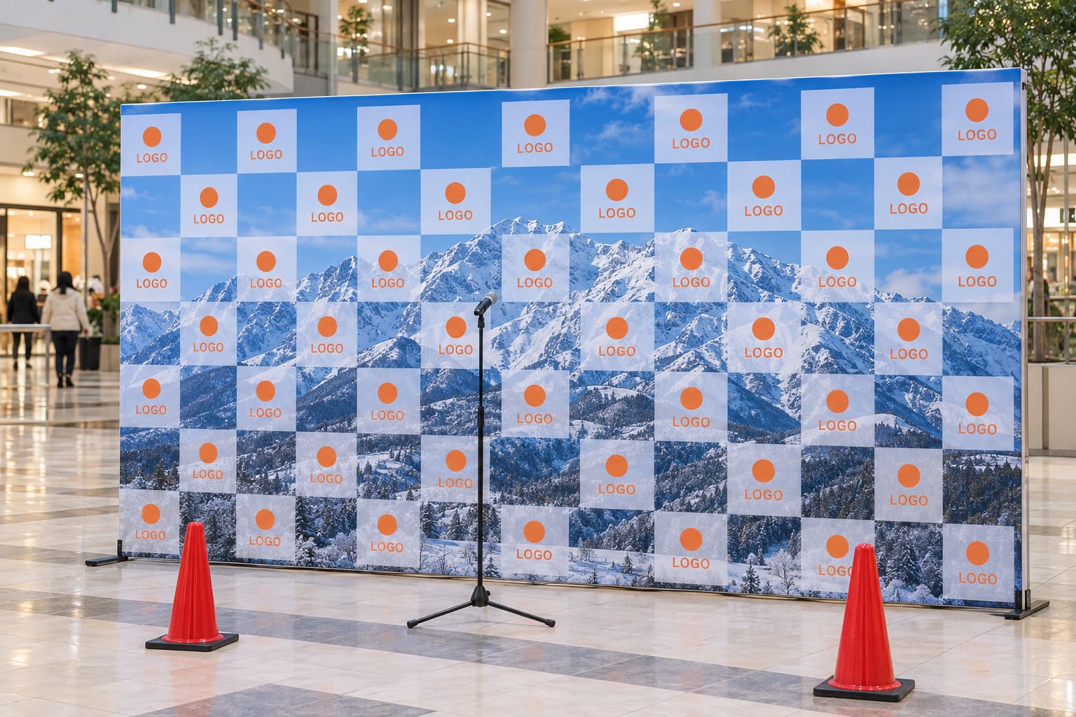 青空と山の写真の上にロゴの格子を重ねた横長のバックパネルが、商業施設の広場に立っている様子
