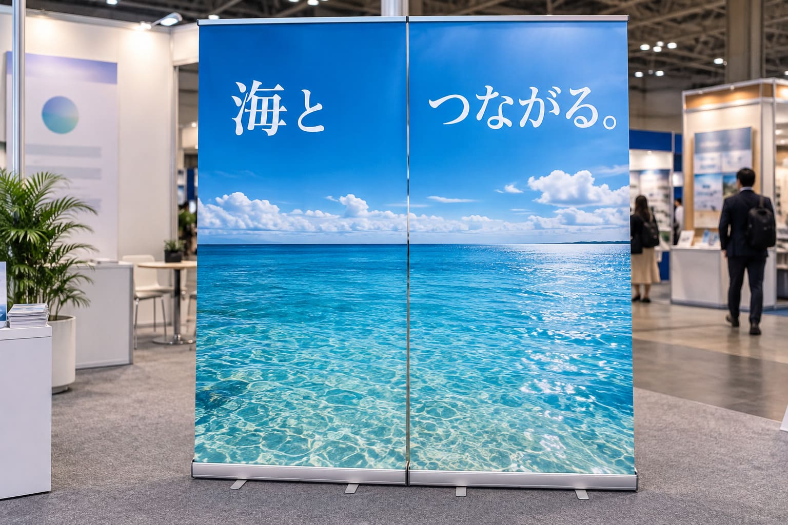 展示会のブースで、2台のロールアップバナーが横に並び、2台で1枚の大きな海と空の写真がつながって見え、上に白い大きな文字のキャッチが入っている様子