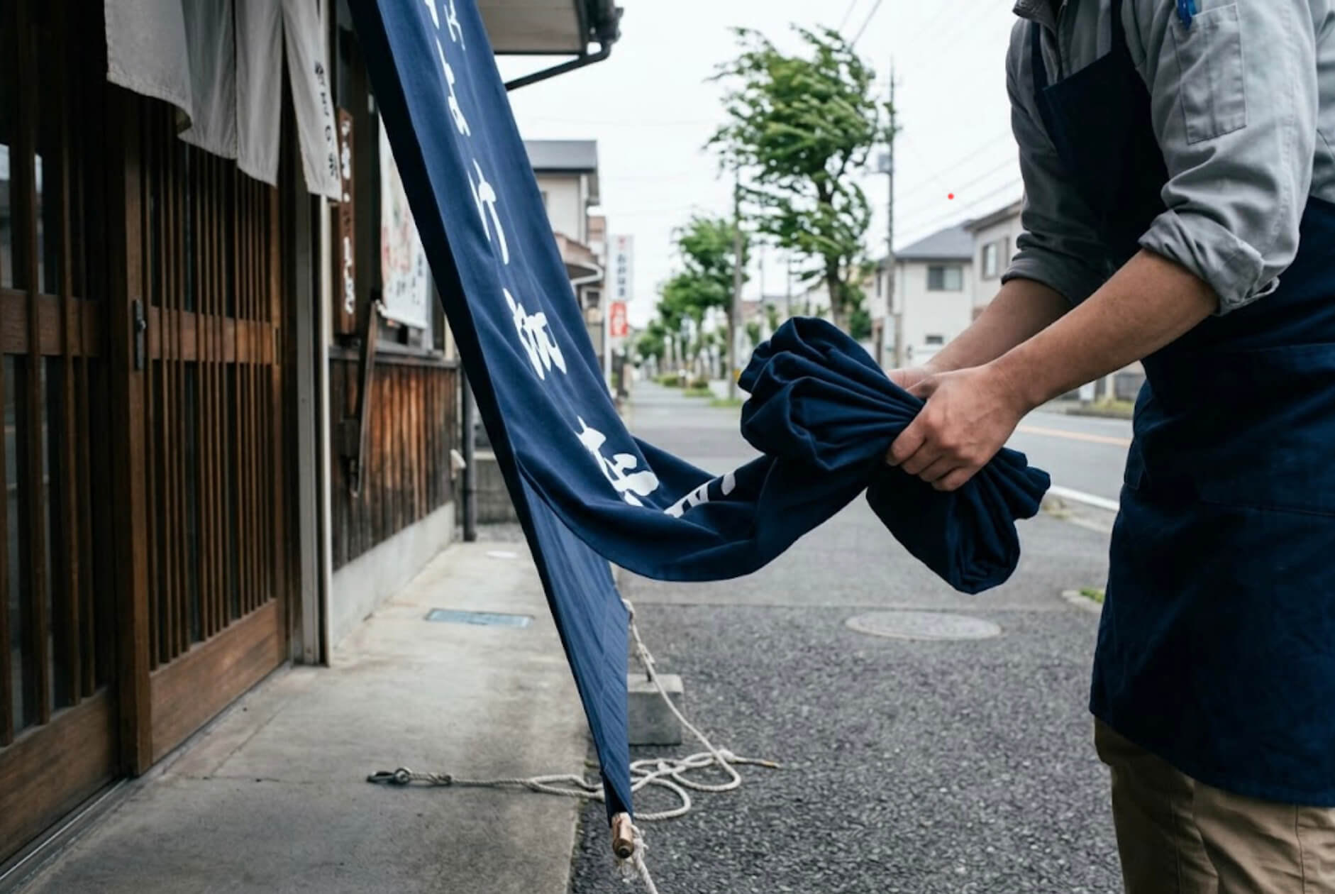 台風の前に、店員が軒先から外した縦長の幕を丸めてしまおうとしている手元の様子