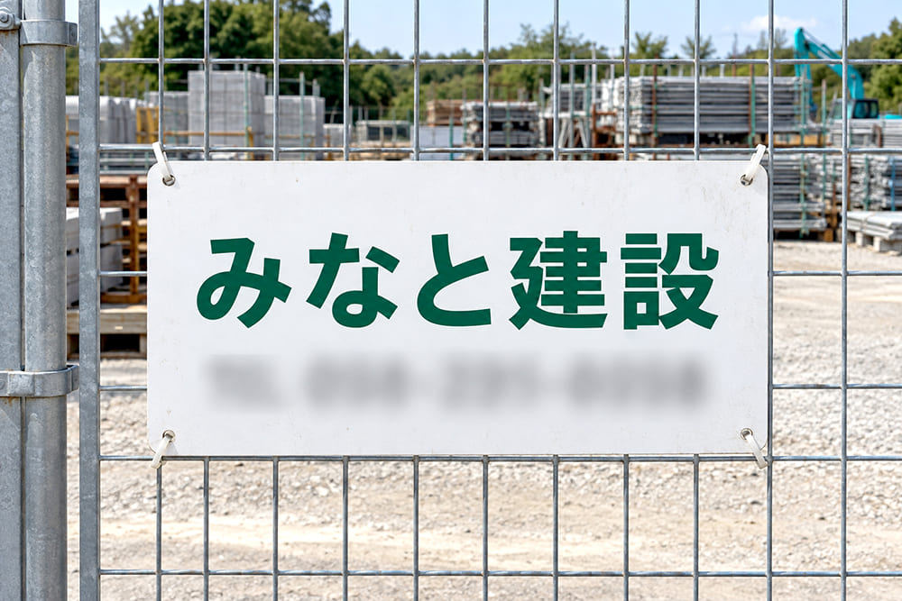 資材置き場のメッシュフェンスに、会社名と電話番号を印刷したプレート看板が四隅で留められている様子（電話番号はぼかす）