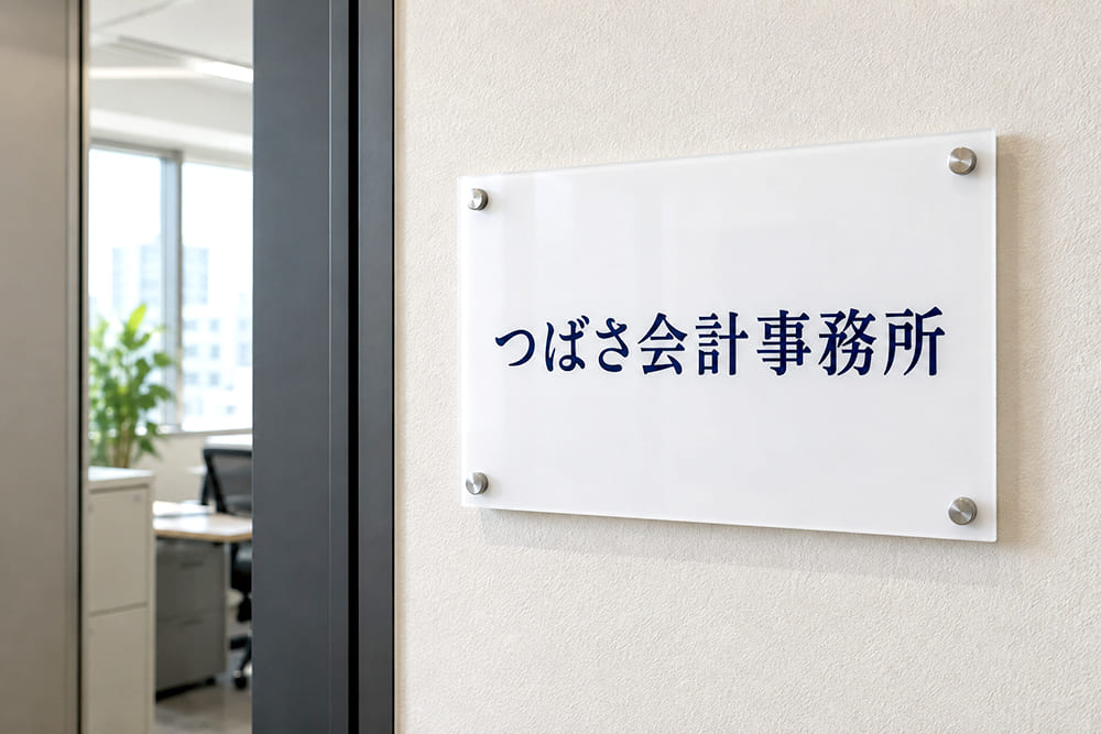 ビルの廊下にある事務所の扉の横に、白いアクリル板に紺色の文字で事務所名が入った小さな看板が付いている様子