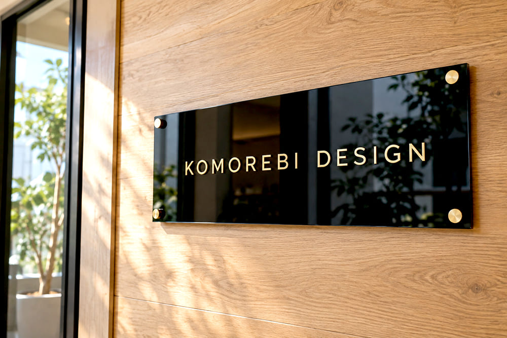 明るい木目の壁に、光沢のある黒いアクリル板に金色の文字で会社名が入った看板が、化粧ビスで取り付けられている様子