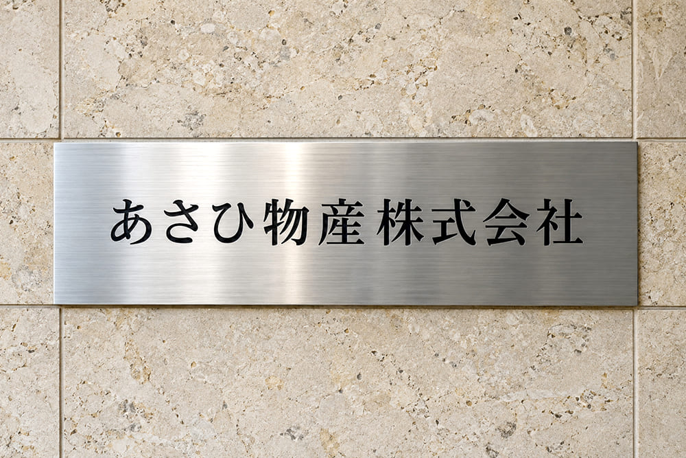 オフィスビルの入口の石張りの壁に、ヘアライン仕上げのステンレス板に黒い文字で社名が入ったエッチング銘板が付いている様子