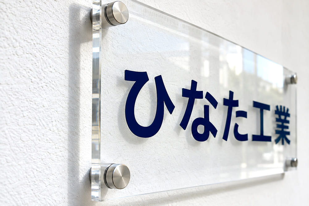 白い壁に、透明のアクリル板の社名看板が四隅の化粧ビスで壁から浮かせて付けられ、壁に板の影が落ちているのを斜め横から写したところ