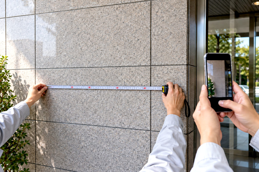 会社の入口の壁の前で、作業者がメジャーで看板を付ける範囲の幅を測り、もう一人がスマートフォンで壁全体を撮っている手元