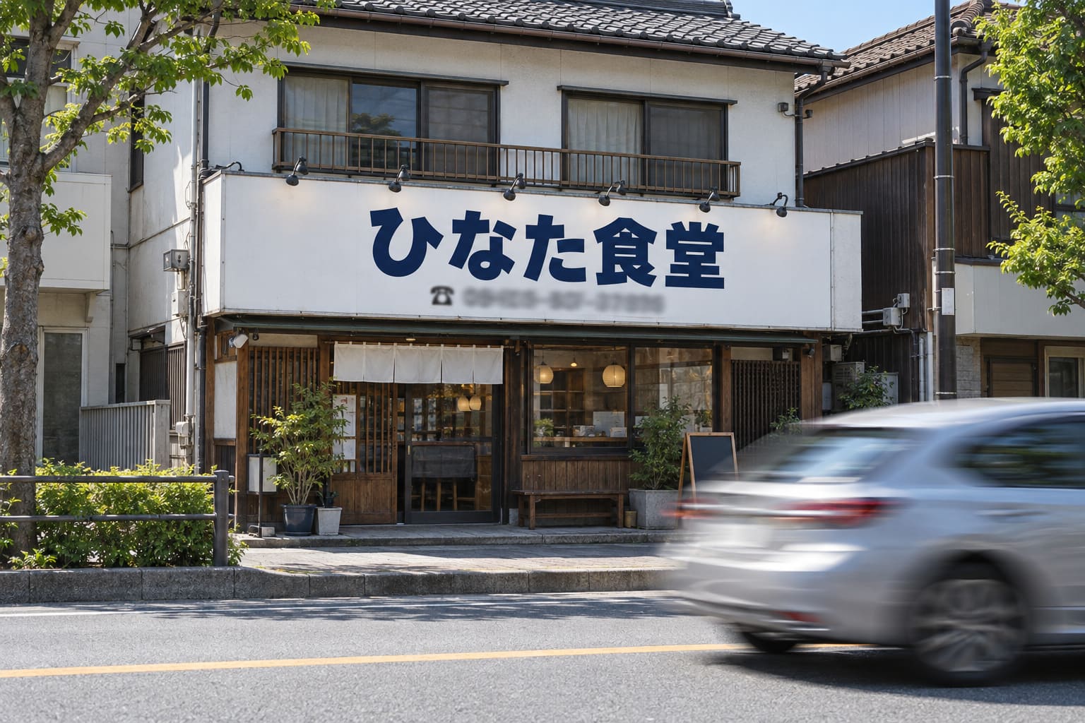 道路の向かい側の歩道から撮った、白い壁面看板に紺色の太い角ゴシック体で「ひなた食堂」と書かれた2階建ての店舗の外観