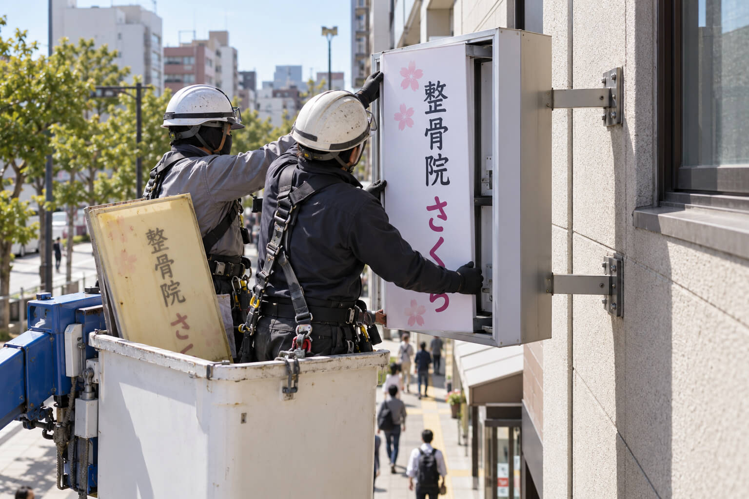 高所作業車の上で、職人が袖看板の枠に新しい面板をはめ込んでいる様子