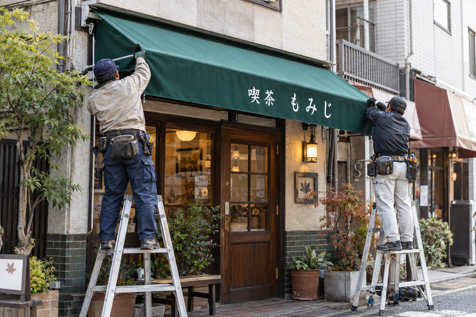 店舗の軒先で、職人がテント看板のフレームに新しい生地を張っている様子