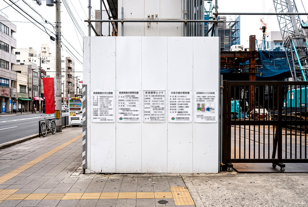 工事現場の仮囲いの出入口付近に、白い標識板が何枚か並んで掲げられている様子