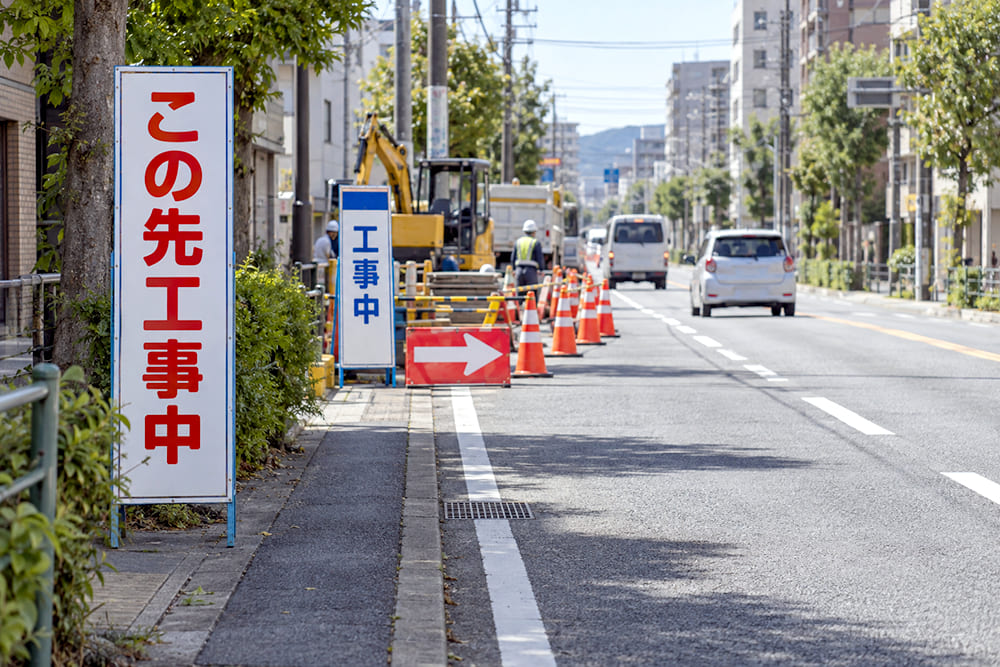 片側の車線を規制している市街地の道路工事の現場で、手前から工事予告の立て看板、赤い矢印板、オレンジのコーンの列、白地に青い帯の工事の看板が並んでいる様子
