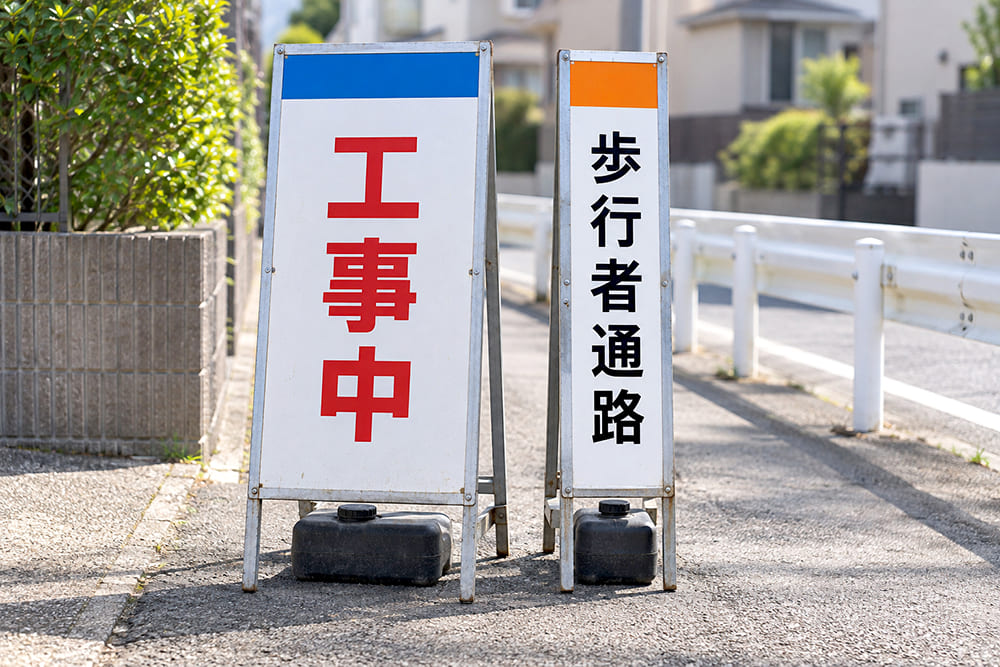 歩道の脇に、幅の広いSL立看板「工事中」と、幅が半分の細いスリムの立看板「歩行者通路」が並んで立っている様子