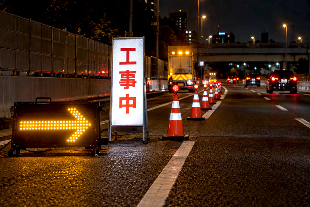 夜の道路工事の現場で、黄色く光るLEDの矢印板が左の車線へ寄るよう示し、その奥に反射して光る工事看板とコーンが並んでいる様子