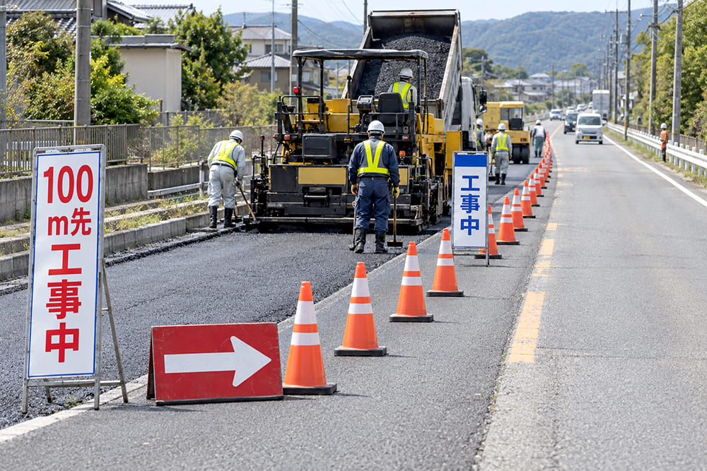 片側の車線を規制している道路工事の現場で、手前から予告の看板、LEDの矢印板、オレンジのコーンの列、工事中の看板が並んでいる様子
