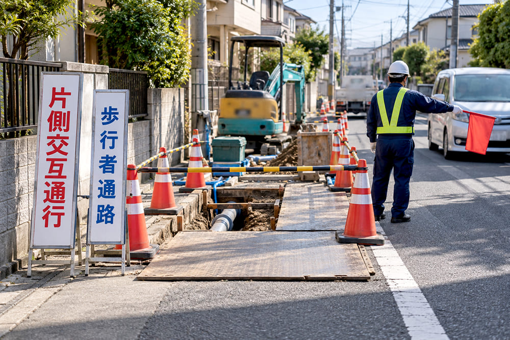住宅街の道路で水道管の工事をしていて、掘削した部分がバリケードで囲まれ、歩行者通路の看板と片側交互通行の看板が立っている様子