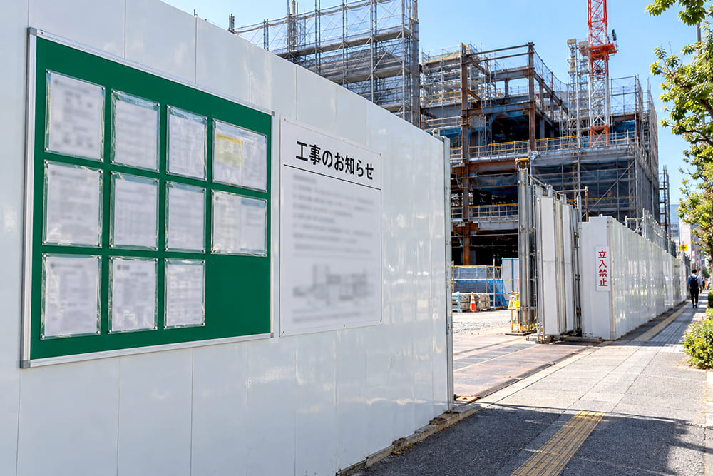 白い仮囲いで囲まれた建築現場のゲートの横に、法令の掲示物が並ぶ掲示板と工事のお知らせ看板、立入禁止のマンガ標識が掲げられている様子