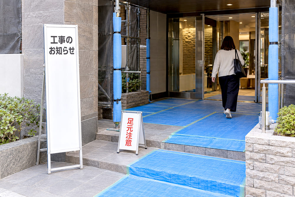 マンションのエントランス付近で改修工事をしていて、床が養生され、工事のお知らせの看板と足元注意の小さな看板が置かれている様子