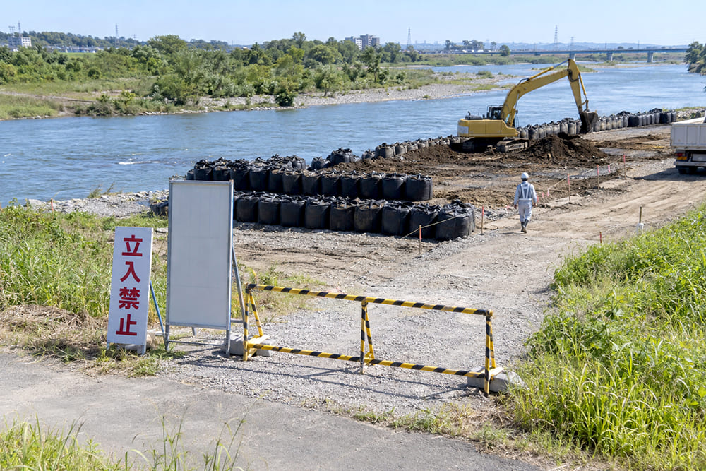 河川敷の工事現場の入口に立入禁止の看板とバリケードが置かれ、奥に大型土のうと重機が見える様子