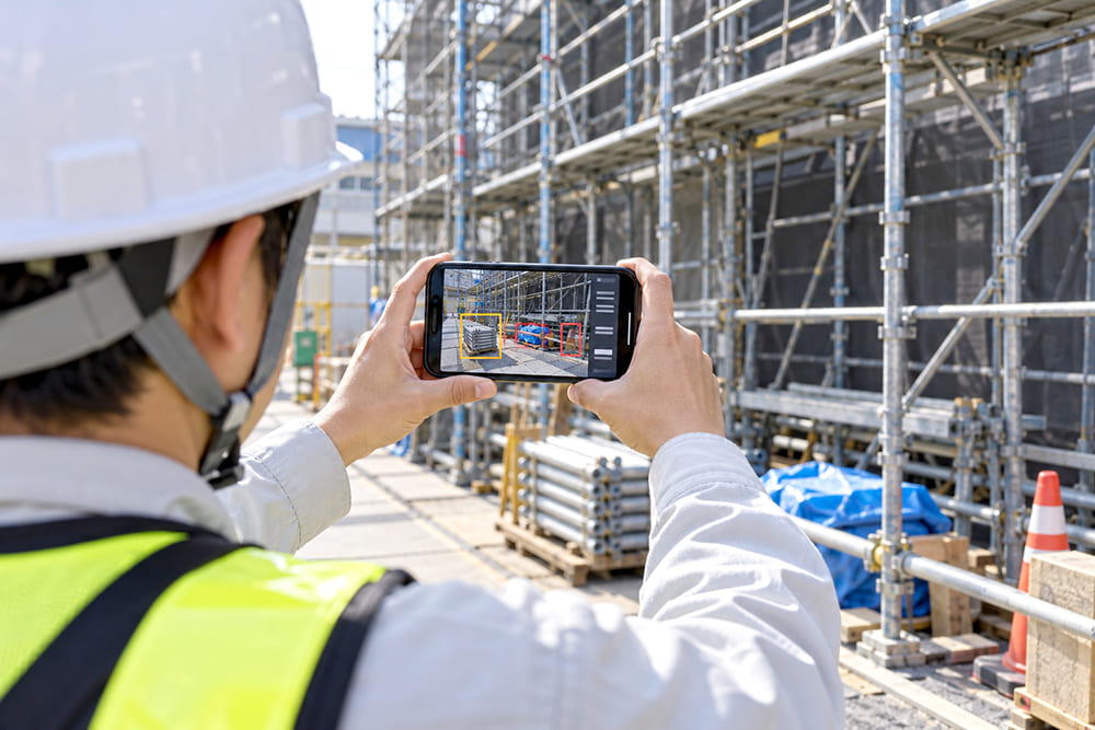 工事現場で、ヘルメットをかぶった現場監督がスマートフォンで足場の周りを撮影していて、画面に危険の候補を示す枠が表示されている様子