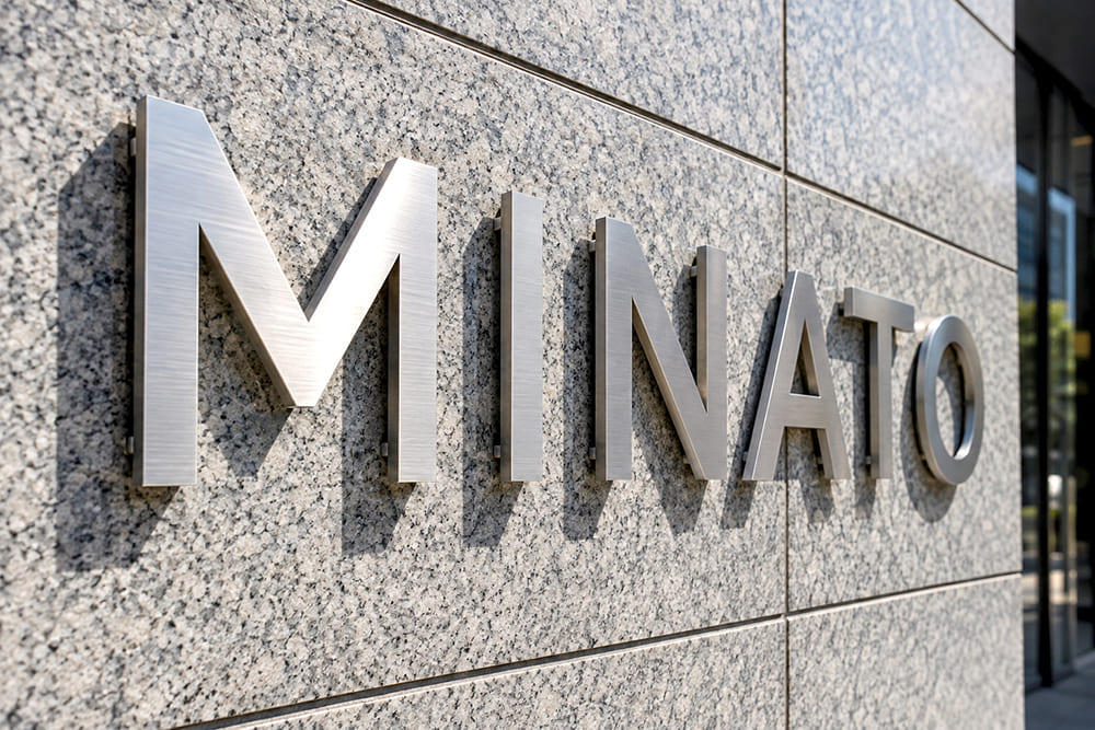 オフィスビルの1階の石張りの外壁に、ヘアライン仕上げのステンレスの切り文字で架空の社名「MINATO」が壁から少し浮かせて取り付けられ、文字の影が壁に落ちている様子