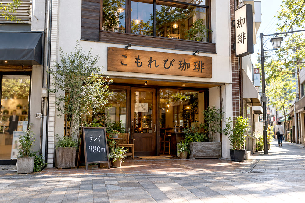 昼の商店街の路面店。正面の壁に架空の店名の横長の看板、2階の壁から縦長の突き出し看板が出て、入口の前の敷地にA型のスタンド看板が置かれている様子