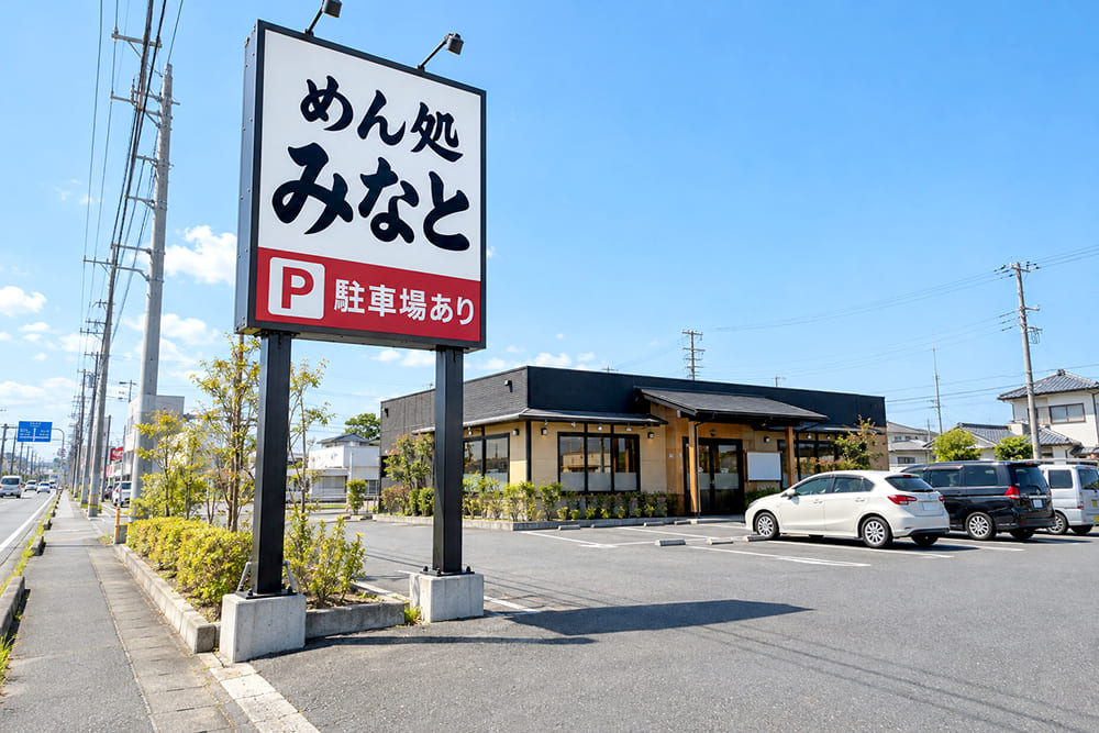 郊外の幹線道路沿いの店の敷地の入口に、背の高いポール看板が立ち、架空の店名と「P 駐車場」の表示が出ている様子