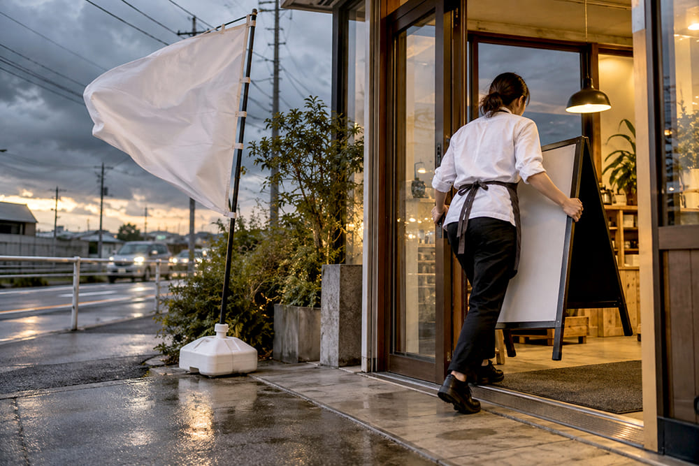 台風が近づく夕方、店の人が入口の前のA型スタンド看板とのぼりを店の中にしまっている様子