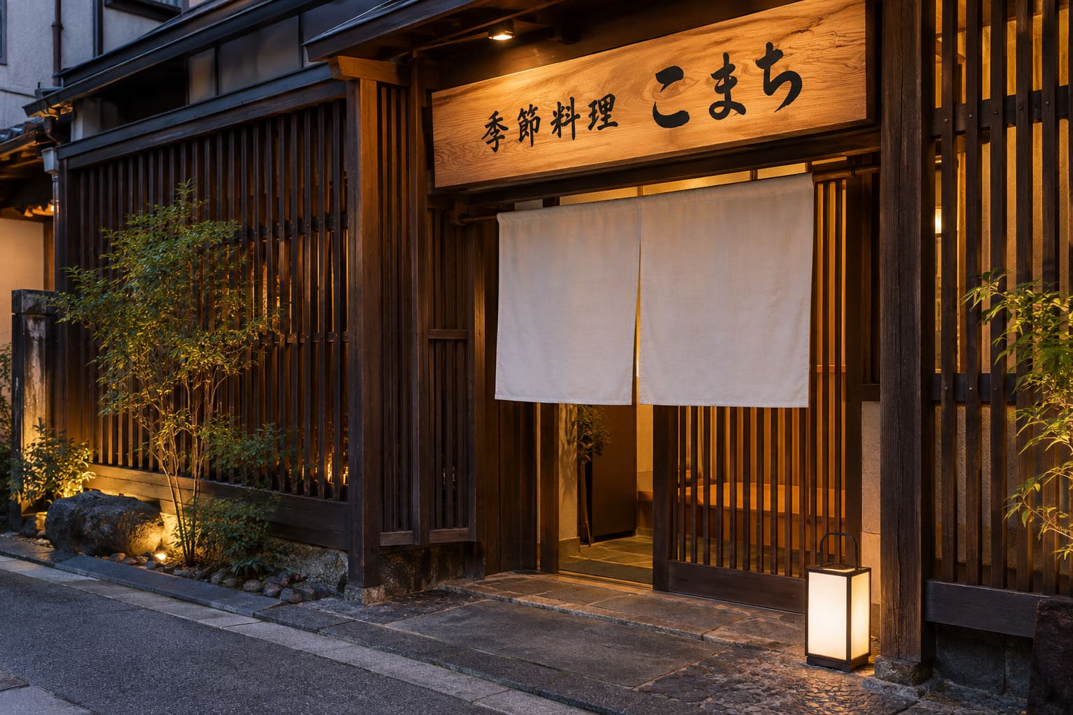 濃い茶色の格子の壁の和食店の入口の上に、杉板に墨色の文字で「季節料理 こまち」と書かれた看板と、白いのれん