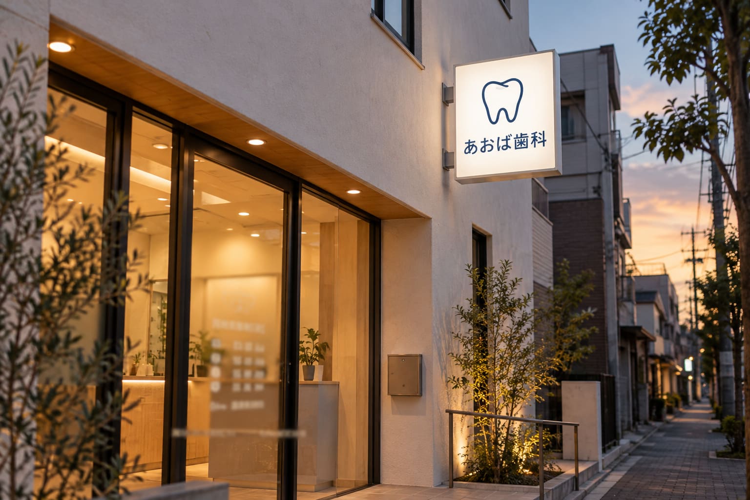 白い外壁のクリニックの2階から突き出した、白地に紺色の歯のマークと「あおば歯科」の文字の四角い看板が夕方に光っている様子