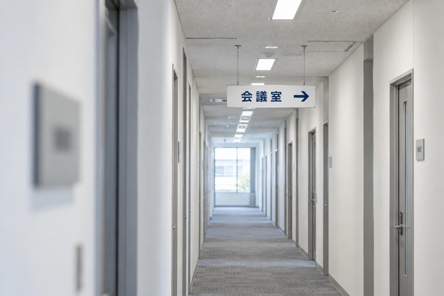 オフィスの廊下で、壁の室名札と天井から吊り下げた案内サインを、離れた場所から見た様子