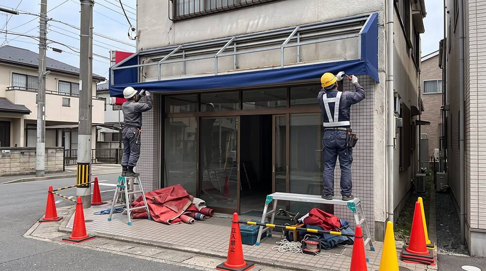 作業着の職人2人が、店舗の入口の上で、既存のフレームに新しいテント生地を張っている後ろ姿